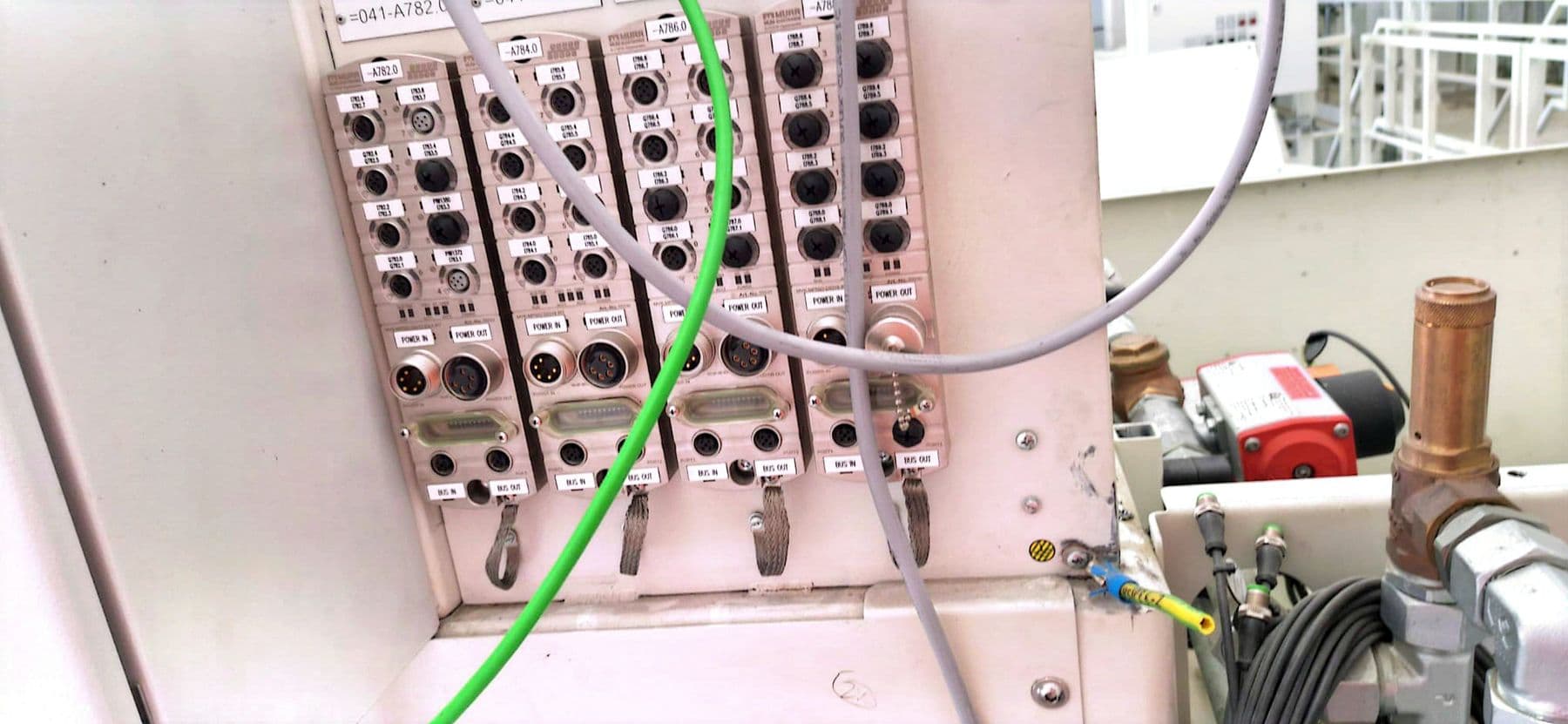 Vier Feldbusmodule an einer Anlagenwand mit Spannungs- und Busanschlüssen, davor ein grünes und ein graues Kabel